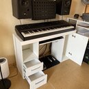 Černý digitální klavír umístěný na bílém stole s přihrádkami a šuplíky. Nad klavírem jsou umístěny dva černé studiové monitory a akustický panel. Vpr…
