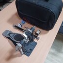 Na fotografii je černý bubnový pedál s kovovými mechanismy a šedým stupněm. Na stupni je viditelné logo Pearl. Vedle pedálu leží černá přepravní tašk…
