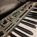 Šedý synthesizér s klaviaturaí. Na ovládacím panelu jsou viditelné nápisy VCO, SOURCE WAVE a GLIDE. V pravém horním rohu je částečně viditelný nápis…