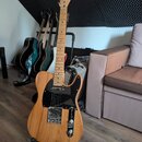 Elektrická kytara s přírodním dřevěným povrchem, černým pickguardem a chromovanými komponenty. Na hlavě je viditelné logo "Telecaster". V pozadí stoj…