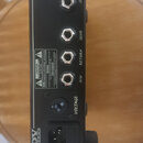 Zadní strana černého zesilovače značky DV MARK. Jsou zde vidět konektory s nápisy SPEAKER OUT, SEND, RETURN a FSW, přepínač VOLTAGE (120/230) a vstup…