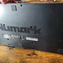 Spodní strana černého elektronického zařízení s velkým nápisem Numark uprostřed. Na spodní části je vidět nálepka s varovnými texty, piktogramy a čár…