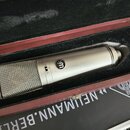 Kondenzátorový mikrofon stříbrné barvy v černé polstrované přepravní kufře. Na těle mikrofonu je viditelné logo „m“ v kroužku a přepínač s označením…