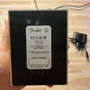 Zadní strana černého kvádrovitého zařízení s kovovými zlatými hranami. Na středu je bílá nálepka s logem Fender a nápisy: MADE IN CHINA, TYPE: PR5296…
