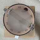 Fotografie zobrazuje snare drum (malý buben) s kovovým obručím a pleteným mechanismem na straně. Na vnitřní straně obruče je vidět černé logo s nápis…