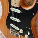 Elektrická kytara s přírodním dřevěným povrchem těla a černým pickguardem. Nástroj má tři bílé snímače typu single-coil, hřebenový kobylník a tři ovl…
