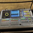 Digitální mixovací pult s šedým povrchem, vybavený dotykovou displejem a několika sekcemi posuvníků. Na pravé straně jsou viditelné barevná tlačítka…