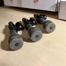 Tři dynamické mikrofony s kovovými mřížkami a tmavě šedým tělem, umístěné v držácích. Na čele mřížky každého mikrofonu je viditelné kulaté logo s pís…