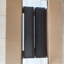 Dvě černé, vysoké a úzké reproduktory umístěné v původním kartónovém balení s bílým polystyrenem. Povrch reproduktorů je matný a v přední části mají…