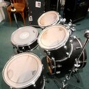 Černá bubnová sada s kovovým hardwarem. Na blbách jsou viditelné loga značky REMO. Sada obsahuje basový buben, dva tom-tomy a jeden snare drum. V poz…