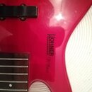 Detail červené elektrické basové kytary s hnedým hmatníkem. Na těle je černý nápis PROFESSIONAL HOHNER a pod ním stylizované B Bass. Povrch laku vyka…