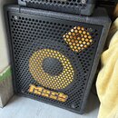 Černý basový reproduktor s kovovou mřížkou, na které je žlutý kruh a čtverec. V dolní části mřížky je umístěno žluté logo Mark bass. Nad ním je vidět…