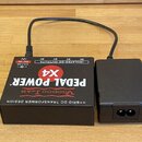 Černý napájecí adaptér a připojená k němu černá krabička s nápisy Voodoo Lab, PEDAL POWER X4 a HYBRID DC TRANSFORMER DESIGN. Na horní straně krabičky…