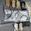 Sada komponentů pro elektrickou kytaru rozložená na šedé perforované podložce. Jsou zde pět zlatých snímačů, černý plastový rám a několik součástek v…