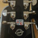 Záběr na hlavu černé kytary s chromovanými ladičkami. Na zadní straně hlavy je zlatý nápis 10021509243 a v dolní části bílé logo Epiphone s nápisy LI…