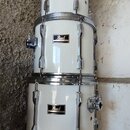 Sada bílých bubnů značky Pearl s chromovaným kováním. Na každém z viditelných bubnů je umístěno logo Pearl v černé rámečce. Horní buben má na obruchu…