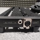 Černý kovový přístroj s dvěma vstupy na zadní straně. Jsou zde nápisy GUITAR IN a LAMP, mezi kterými se nachází jeden 6,3mm jackový vstup a jeden XLR…