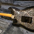 Elektrická kytara v černém lakovaném provedení s ornamentálním vzorem paisley na zadní straně těla. Kytara leží v šedém plyšovém kufru, viditelný je…