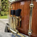 Detailní záběr na dřevěný buben s zlatým kováním. Na korpusu je připevněn zlatý držák s dvěma černými plastovými knoflíky. Povrch kování vykazuje dro…