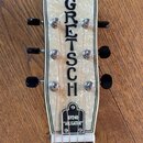 Hlava kytary s bílým perleťovým povrchem a černým lemováním. Uprostřed je svisle napsáno logo GRETSCH a v dolní části nápis G9240 "ALLIGATOR". Nástro…