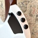 Detail zadní strany bílé elektrické kytary s hnědým krkem. Na těle jsou tři černé oválné potenciometry a černý plastový kryt elektroniky. Na kytáře n…