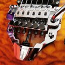 Detail chromovaného tremolo systému na kytáře s oranžovo-hnědým plamenovitým povrchem. Na kovové části je v dolní části vyryté logo „ Floyd Rose“ a p…