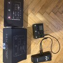 Na dřevěné podlaze jsou dvě černé krabice a dva přístroje. Menší černý efektový pedál má nápis Line 6 HX STOMP a připojení k černému zařízení s bílým…