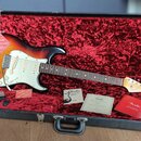 Elektrická kytara s tělem v barvě sunburst a bílým pickguardem leží v otevřeném černém kufru s červeným plyšovým vnitřkem. Na hlavě kytary je viditel…