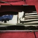 Černý gramofon s lesklým povrchem, v němž je vidět odraz okolí. Na přední straně je nápis HI FI TURNTABLE a vedle něj nečitelný kód. V pravé dolní čá…