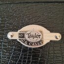Detail kovového štítku na černém kufru s texturou kůže. Štítek obsahuje logo Taylor a nápisy QUALITY GUITARS & CASES. Je připevněn dvěma šrouby, povr…