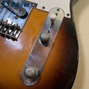 Detail elektrické kytary s tělem v barvě sunburst a bílým pickguardem. Na těle je připevněna kovová kontrolní plocha s dvěma šedými vrutovými knoflík…