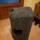 Šedý reproduktor v potahu z hrubé plsti. V přední části je vidět obdélníkový otvor, skrze který je patrný černý vnitřní prvek. Na povrchu plsti nejso…