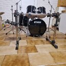 Černá bubnová sada s chromovaným hardwarem. Na basovém bubnu je bílý nápis TAMA a menší, nečitelný nápis v dolní části. Sada obsahuje basový buben, d…
