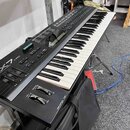 Černý digitální syntezátor s bílými a černými klávesami. Na levé straně je vidět nápis YAMAHA a model DX7. Nástroj obsahuje řadu tlačítek, posuvníků…