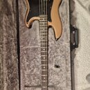 V otevřeném černém kufru s šedým vnitřkem leží elektrická basová kytara. Nástroj má zlatavě hnědý lak, černý pickguard a hnedý hmatník. Na hlavě je v…