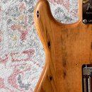 Detail těla elektrické kytary z přírodního dřeva s viditnou masivní strukturou. Na kovovém krytu v pravé části je čitelné logo Fender a nápisy CE a L…