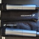Dvě bezdrátová dynamická mikrofony s šedým kovovým tělem a černou mřížkou. Na obou je nápis SENNHEISER XS WIRELESS. Mezi mikrofony jsou dvě černé lát…