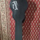 Černý tvrdý kufr na hudební nástroj v kytarovém tvaru. Na horní části je bílý nápis „Gibson GEAR“. Povrch vykazuje drobné odpodřeniny a známky opotře…