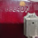 Detail červeného lakovaného povrchu nástroje s kovovým ladicím kolíkem. Na laku je vyražené sériové číslo 886407. Povrch vykazuje známky opotřebení v…
