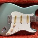 Elektrická kytara s kovově šedým lakem v tónu sage green, umístěná v oranťovém plišovém kufru. Nástroj má bílý pickguard, tři jednoduché snímače s kr…