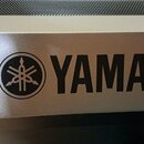 Detail šedého kovového povrchu hudebního zařízení. Na přední straně je černý nápis YAMAHA a vedle něj kulaté logo značky Yamaha. Povrch je hladký s m…
