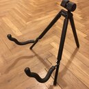 Černý stojan na kytaru s třemi nohama a dvěma zakřivenými opěrkami. Na horní části je viditelný nápis Spider-EA. Stojan stojí na dřevěné podlaze v ry…