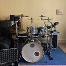 Černá bicí souprava s bílým předním zrcadlem basového bubnu, na kterém je logo MAPEX. Na stojanu je viditelný nápis Gibraltar. Souprava obsahuje něko…