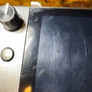 Detailní záběr na horní část elektronického zařízení se stříbrným šasi a černým displejem. Na levé straně je vidět černý otočný knoflík a jedno kulat…