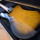 Zadní strana elektrické kytary s tělem z dřeva v žlutomodrém přechodovém lakování (sunburst), umístěná v černém tvrdém kufru. Na těle jsou vidět šest…