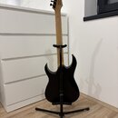 Černá elektrická kytara s jasným dřevěným hmatníkem, umístěná na černém stojanu. Na hlavě kytary je viditelné logo "Ibanez". Tělo nástroje má černý p…