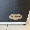 Detail černého přepravního kufru s texturovaným povrchem a hliníkovou hranou. Na středu je oválný kovový štítek s nápisem ROCKCASE® by warwick®. Povr…