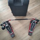 Červený dvojpedál na bicí buben s kovovými šlapkami a černým spojovacím hřídelem. Na obou pedálech je nápis „The Drummer's Choice“ a na pravém pedálu…