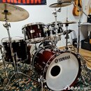 Tmavě hnědá bicí souprava s kovovým hardwarem. Na basovém bubnu je viditelné logo a nápis YAMAHA, v pozadí visí vlajka s nápisem AMAHA. Souprava obsa…