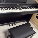 Černé digitální piano s doprovodnou černou polstrovanou stoličkou. Na horní části nástroje je umístěn nápis YAMAHA. Pod klávesnicí jsou vidět tři sus…